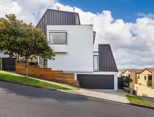 Street-level exterior photograph of a contemporary multi-level townhouse or residential home on a sloping suburban site. The facade combines white horizontal weatherboard cladding with dark grey vertical metal cladding and a dark double garage door. A timber privacy fence, landscaped front garden, driveway, and large projecting windows are visible. The pitched and angled roof forms create a modern composition, with neighbouring attached homes in the background under a bright blue sky.