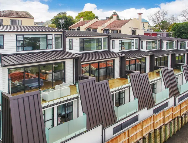 Exterior aerial view of a contemporary multi-unit residential development with repeated townhouse-like dwellings, dark metal pitched roof sections, white horizontal cladding, expansive glazing, private balconies with glass balustrades, and timber privacy screens. Established neighbouring houses and mature trees are visible in the background.