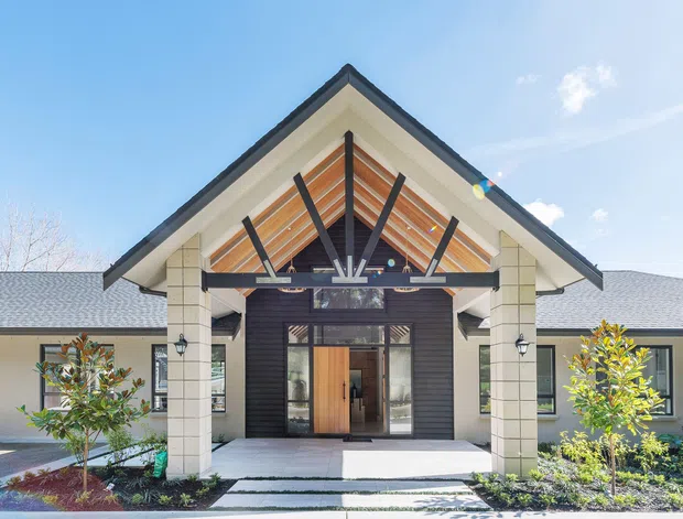 Front exterior view of a single-storey residence with a prominent gabled entry porch, white rendered masonry columns, dark vertical cladding around the entrance, exposed timber rafters and a glazed timber front door. Symmetrical wings extend to either side, with landscaped garden beds and a paved approach.