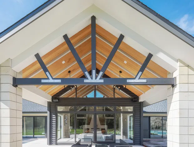 Exterior architectural view of a contemporary entrance beneath a large gabled canopy. The facade combines pale rectangular masonry cladding, dark metal framing, extensive glazing, and a warm timber-lined vaulted soffit with exposed structural beams. Interior spaces are visible through the glazed entry, with landscaped grounds and adjacent roof forms in the background.