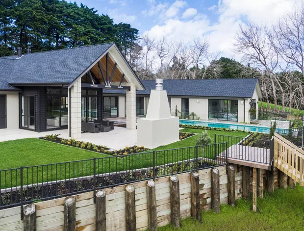 Exterior view of a contemporary single-storey home with a steep gabled roof, light masonry walls, dark window frames, and extensive glazing. The landscaped grounds include a manicured lawn, paved terrace, raised timber retaining wall, black metal fencing, an outdoor fireplace or chimney structure, and a rectangular swimming pool beside the house.