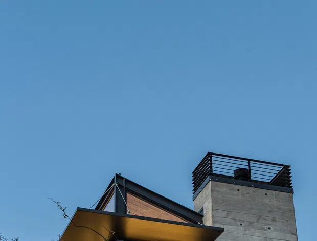 A low-angle exterior view of a contemporary multi-level house against a clear blue sky. The building features a projecting flat-roofed canopy, dark metal framing and balcony railings, warm timber wall cladding, and a tall exposed concrete tower with a rooftop terrace. Leafless vegetation partially obscures the lower facade.