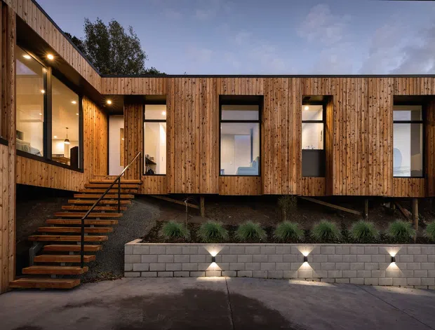 Twilight exterior view of a contemporary multi-level house clad in vertical natural timber, with large dark-framed windows, a projecting upper volume, open-riser timber stairs, and a landscaped retaining wall with integrated uplighting. Warm interior lighting contrasts with the cool evening sky.