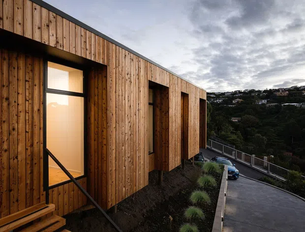 Exterior view of a contemporary elongated building clad in vertical timber boards, with deep-set windows and projecting timber-framed openings. The building sits on a sloped, landscaped site above a paved driveway, with a suburban hillside visible beyond under an overcast sky.