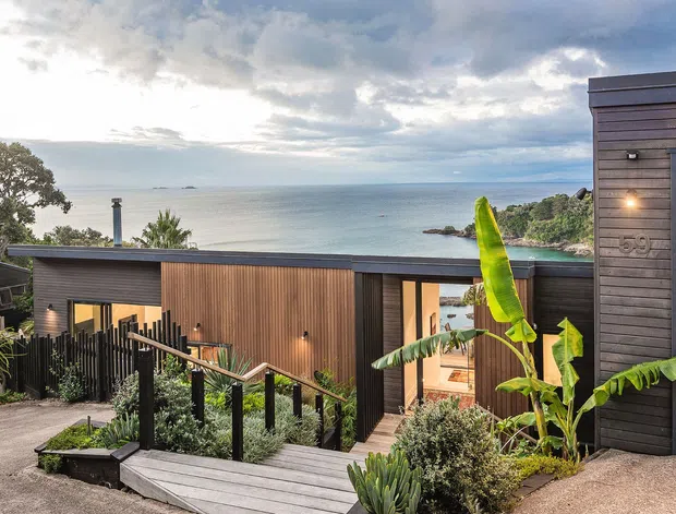 A contemporary coastal home viewed from an elevated outdoor approach, featuring dark horizontal cladding, warm vertical timber screening, a flat roofline, and a landscaped pathway with tropical planting. The dwelling overlooks a calm ocean bay and rocky shoreline.