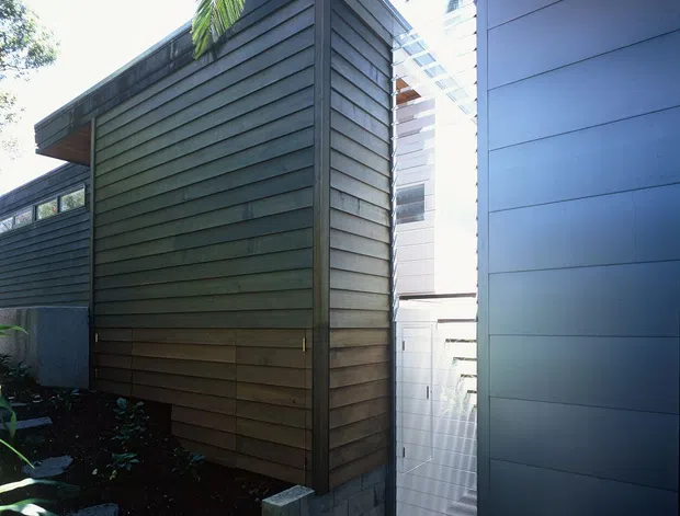 A close exterior view of a contemporary multi-volume building, showing dark horizontal timber weatherboard cladding, a contrasting smooth blue-grey cladded wall, a narrow glazed connection or passage, and tropical palm vegetation around the structure.