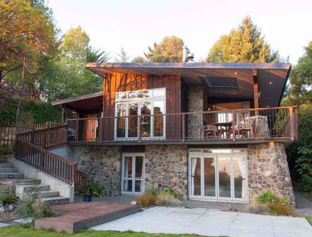 A compact multi-level residential home set among mature trees, with a dark low-pitched roof, warm timber cladding, extensive glazing, and a lower level faced in irregular natural stone. A raised timber deck with railing fronts the upper living level, while stairs, paved areas, and a timber walkway connect the split-level exterior.