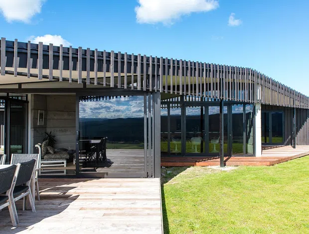 Contemporary single-storey building with extensive floor-to-ceiling glazing, dark framed windows, and a distinctive curved facade wrapped in closely spaced vertical timber-look screening. A broad timber deck with outdoor dining and lounge furniture fronts the building, opening onto a manicured lawn. The reflective glazing captures surrounding hills and sky.