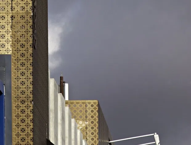 Upward-looking exterior architectural photograph showing tall urban building facades under a dark overcast sky. The prominent facade is clad in repeating yellow-gold patterned tile or panel work with dark geometric detailing, alongside a dark textured wall and vertical pale concrete elements. A projecting black structural canopy or beam and modern external signage supports are visible. The composition emphasizes the building envelope and strong vertical forms.