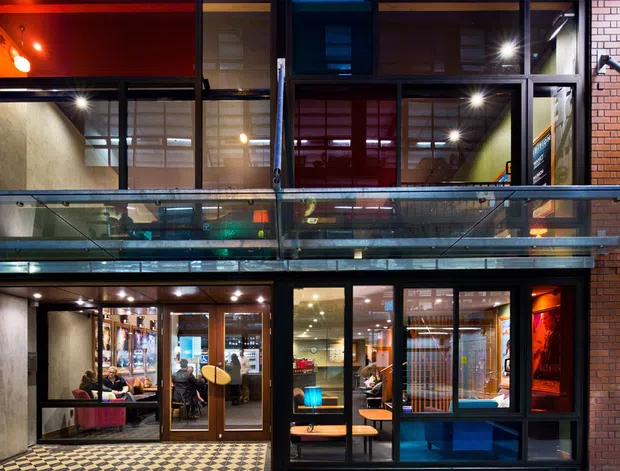 Night-time exterior view of a multi-level urban commercial building, with exposed red brick walls, dark metal-framed glazing, illuminated interiors, and a projecting glass-and-metal balcony or walkway. The ground floor appears to contain hospitality or retail seating, while upper-level spaces are visible through windows. Strong contrast between warm interior lighting and dark blue night tones creates an industrial contemporary character.