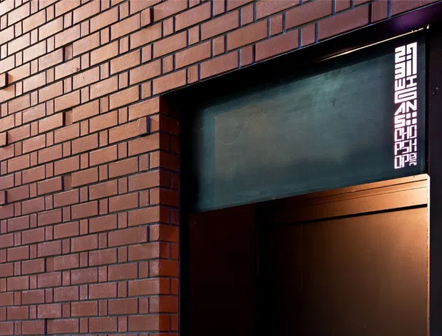 Close-up of a dark red brick building facade with a recessed commercial doorway or entry opening. The entrance has black metal framing, a dark transom panel, warm interior lighting, and a vertical white graphic sign along the right side. The image is tightly cropped to the exterior entry and does not reveal the interior space.