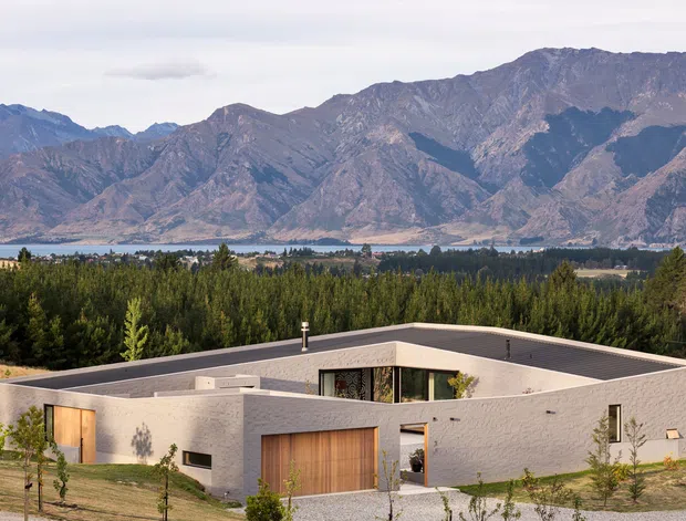 A low, single-storey modern residence with a flat roof and pale textured rendered walls, set in a landscaped rural site. Timber garage and entry doors punctuate the minimalist facade, while large glazing opens toward a dramatic mountain range and lake or valley landscape in the background.