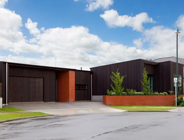 Street-level exterior view of a contemporary low-rise building with dark vertical cladding, red brick feature walls, and large integrated garage doors. The property sits along a suburban street with landscaped front boundaries, concrete driveways, and neighbouring homes visible to the right under a bright partly cloudy sky.