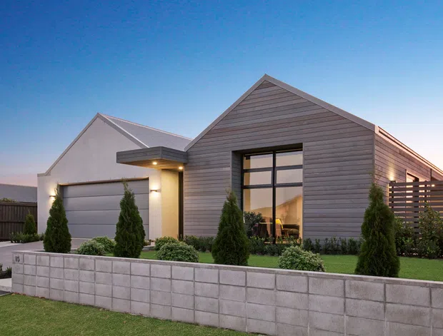 A contemporary single-storey residential home viewed from the front at dusk. The facade combines horizontal grey timber-style cladding with a white rendered garage wing, a steep gabled roof, large black-framed glazing, landscaped planting, a low concrete block retaining wall, and a slatted privacy screen.