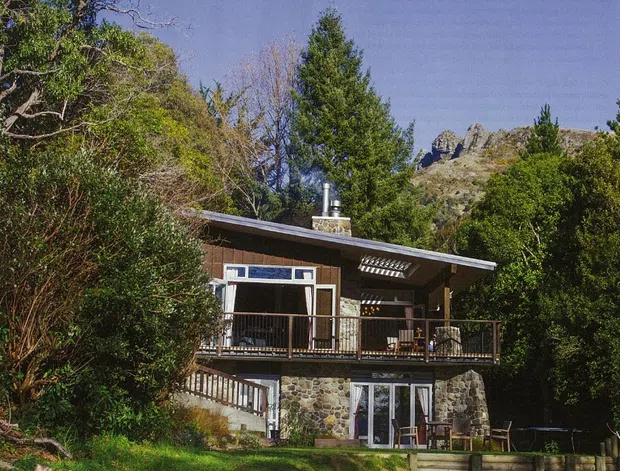 A compact two-storey mountain home nestled among dense vegetation, with a low-pitched roof, timber-clad upper level, stone lower-level walls, broad glazed doors and windows, and a timber-railed balcony. Rocky alpine terrain rises behind the house.