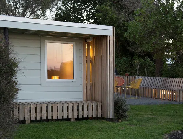 Exterior view of a compact, single-storey timber-clad structure in a landscaped garden. The building has pale horizontal weatherboards, a simple low-pitched or flat roof with white fascia, a small window, and a vertical timber-screened recessed outdoor area. A timber slat deck or balustrade extends around the structure, with a matching fenced patio and outdoor seating visible to the right. Mature vegetation surrounds the scene, and warm interior and exterior lighting create an inviting evening atmosphere.