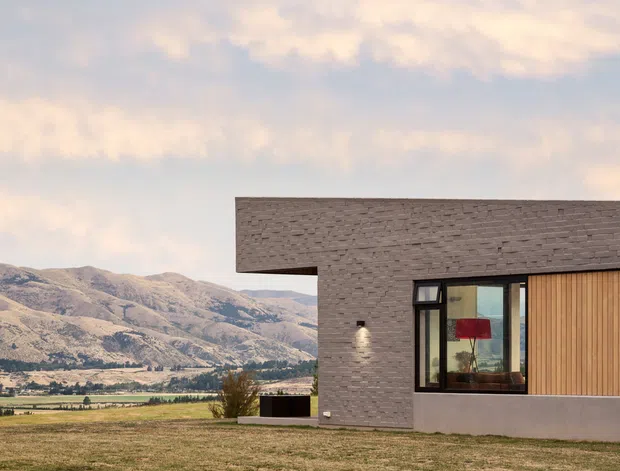 A modern single-storey house exterior set in an open rural landscape, with a low horizontal form, dark textured brick cladding, warm vertical timber wall panels, extensive glazing, a projecting flat roof, and a chimney. Mountains and agricultural fields are visible beyond the property.