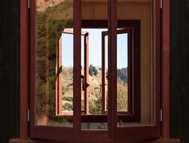 Close-up architectural view of a timber-framed multi-panel window set into dark vertical timber cladding. The deep red-brown timber casements are open inward, revealing layered window openings and a sunlit dry, hilly landscape beyond. The image emphasises the window construction, natural timber surfaces, and strong indoor-outdoor visual connection.