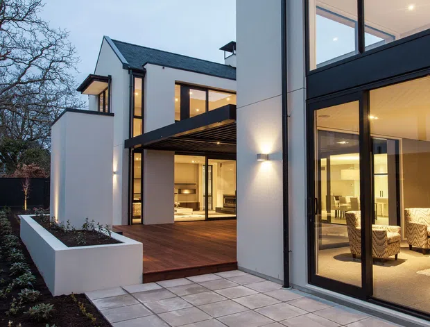 Twilight view of a contemporary multi-level house with crisp white rendered walls, dark roof and window frames, extensive floor-to-ceiling glazing, and a timber-clad covered terrace. A paved courtyard and raised white planter frame the entrance, while warm interior lighting reveals living and dining areas through the glass.