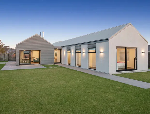 Exterior view of a contemporary single-storey residential dwelling with two connected gabled volumes. The home has pale rendered walls, grey standing-seam metal roofing, horizontal timber-look cladding on the smaller wing, extensive dark-framed glazing and a broad manicured lawn with concrete paths.