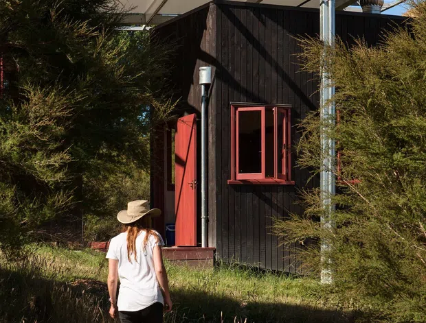 Exterior view of a compact dark-clad cabin or small outbuilding set within dense natural landscaping and tall dry grasses. The building has a simple rectilinear form, red-painted door and window trim, and a broad lightweight flat canopy with exposed metal framing and translucent or pale roof panels. A person approaches the open entrance, providing scale. Bright clear daylight and strong shadows emphasize the rustic bush setting.