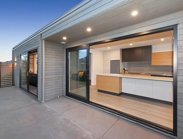 Contemporary single-storey dwelling exterior with a broad covered terrace and fully open sliding glass doors revealing a compact modern kitchen and adjacent interior. The facade uses horizontal light-grey cladding, dark aluminium framing, and a timber-lined soffit with recessed lighting. The kitchen features two-tone timber and white cabinetry, a dark benchtop and splashback, and pale timber flooring. A slatted privacy fence and distant landscape are visible beyond the concrete terrace.