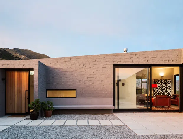 Low, single-storey modern residence with a warm pink-beige masonry facade arranged around a paved courtyard. A tall timber entry door, narrow horizontal window, and expansive black-framed glazed living area face the courtyard, with a glimpse of a richly patterned interior and surrounding rugged mountains beyond.