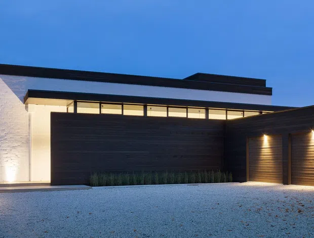 Contemporary single-storey residence viewed from the driveway at dusk. The facade combines a white rendered or masonry wall with dark horizontal timber-like cladding, a low flat roofline, narrow clerestory glazing, recessed warm lighting, and an attached multi-bay garage. A broad pale gravel driveway occupies the foreground.
