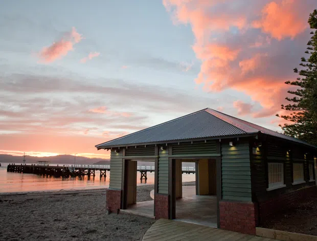 A small waterfront pavilion or boathouse beside a pier, photographed at sunset. The building has green horizontal weatherboard cladding, a dark corrugated pitched roof, brick-clad lower walls and open bays facing the water. Timber decking leads into the structure, with calm water, a pier and distant hills visible beyond.