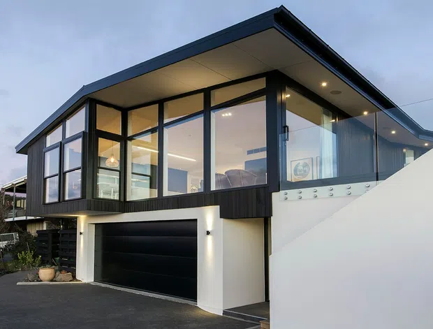 Exterior view of a contemporary two-storey home with a dark low-pitched roof, extensive floor-to-ceiling glazing, dark vertical cladding, and a black garage door. The elevated upper level features a bright open living space behind glass, with a white rendered lower level and a glass-balustraded terrace or balcony. The image is taken from the driveway at dusk.