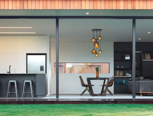 Wide exterior view through floor-to-ceiling glazing of a contemporary single-level home, showing an open-plan kitchen, dining area and living room. The facade has dark metal-framed sliding glass doors beneath a warm timber-lined soffit, with a compact kitchen island and stools on the left, dining table centrally, and a dark living area on the right.