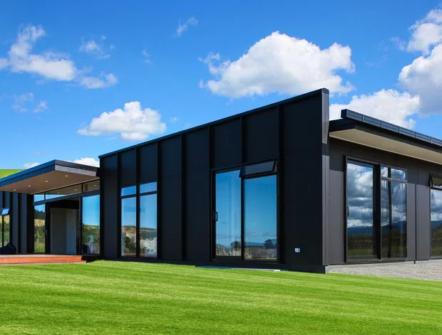 A contemporary single-storey black-clad house set in a broad green rural landscape. The building has strong rectilinear volumes, a low-pitched flat roof, extensive floor-to-ceiling glazing, and a covered outdoor terrace or entry deck. Dark vertical facade panels contrast with the bright lawn and blue sky.
