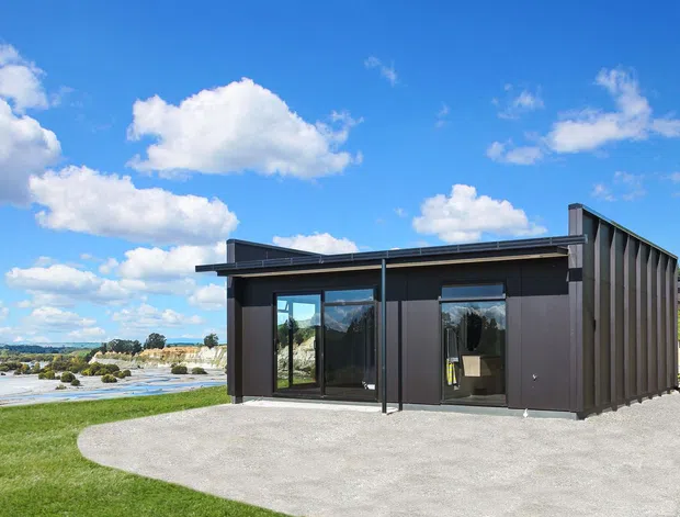 A compact, single-storey contemporary building set beside a broad body of water. The structure has dark ribbed metal cladding, a low mono-pitched roof, large glazed openings, and a simple concrete-paved forecourt. Open lawn, rocky shoreline, and a sunny blue-sky landscape surround the building.