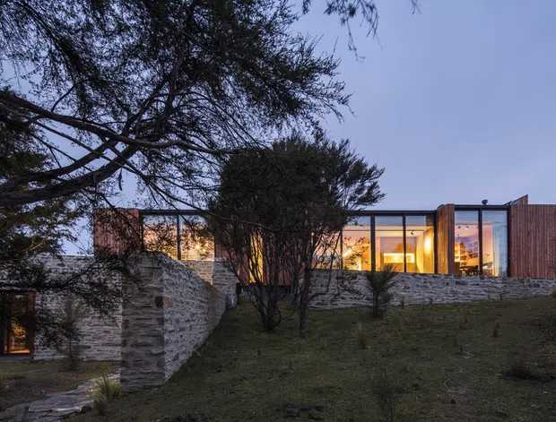 A low, modern house set into a gently sloping landscaped site, photographed at dusk. The building features extensive floor-to-ceiling glazing framed in dark metal, warm timber-clad wall sections, and layered rough-stone retaining walls that integrate the structure into the terrain. Mature trees partially screen the facade, while warm interior lighting creates a welcoming glow.