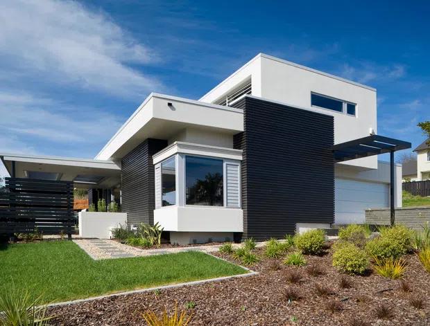 Exterior view of a modern multi-level residence with crisp white rendered walls, extensive dark horizontal cladding, flat roof forms, projecting balconies and roof planes, and broad glazed openings. The home sits within a landscaped suburban garden with lawn, gravel paving, low planting, and a covered carport or entry structure.