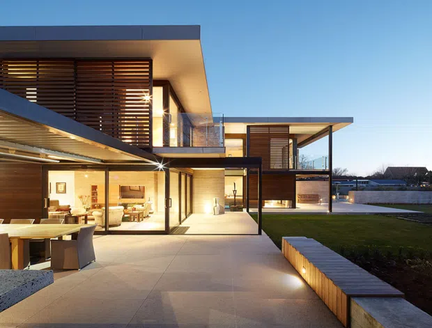 Exterior architectural photograph of a large contemporary multi-level residence at dusk, viewed from a broad paved courtyard or terrace. The house features flat roof planes, extensive floor-to-ceiling glazing, warm timber horizontal screening, dark framed openings, projecting canopies, and layered indoor-outdoor living areas. A manicured lawn, built-in illuminated bench seating, and a glimpse of a swimming pool are visible around the paved forecourt.
