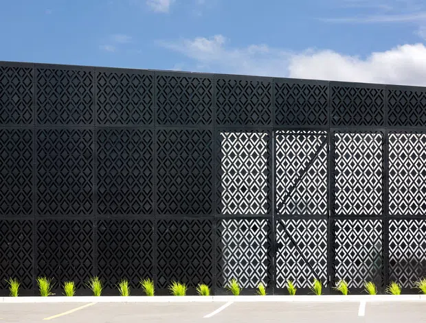 Exterior view of a long, dark facade composed of repeating geometric patterned screening panels. Bright geometric perforations form a contrasting glazed or open screened section, with a narrow planted strip and paved foreground beneath a clear blue sky.