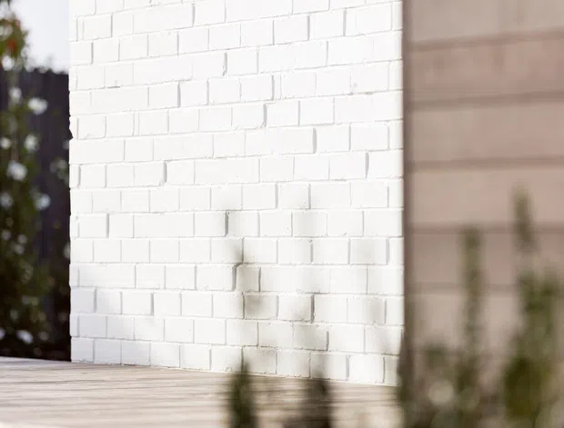 Close-up architectural detail of a white-painted brick wall meeting a pale timber-clad wall, with a light timber deck or platform in the foreground and soft landscaping visible at the edges. The image has a minimalist, contemporary exterior character with shallow depth of field.