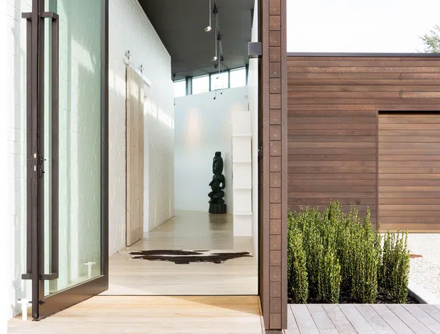 A contemporary residential entry viewed from outside, with a tall glazed pivot door opening into a minimalist hallway. The entrance features pale timber flooring, white tiled walls, a dark sculptural artwork, exposed track lighting, and warm horizontal timber cladding around a recessed garage or service door. A small landscaped planter and timber deck complete the foreground.