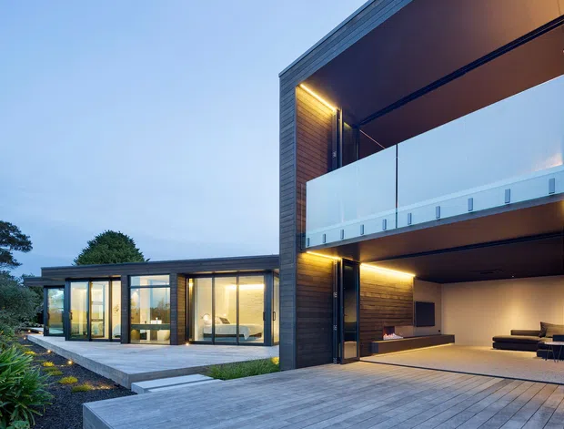 Contemporary multi-level residence at dusk, with dark vertical cladding, extensive glazing, recessed warm lighting, a large open ground-level entertaining area, and a light-toned balcony balustrade. A paved terrace connects the main volume to a low glazed wing, with landscaped planting at the edges.