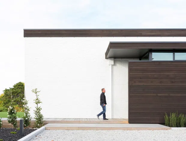 A minimalist contemporary single-storey house exterior with a crisp white masonry facade, dark timber-clad garage door and matching horizontal roof-edge trim. A covered entry walkway and restrained landscaped frontage sit in front of the building, with narrow clerestory glazing beneath the projecting roof canopy.