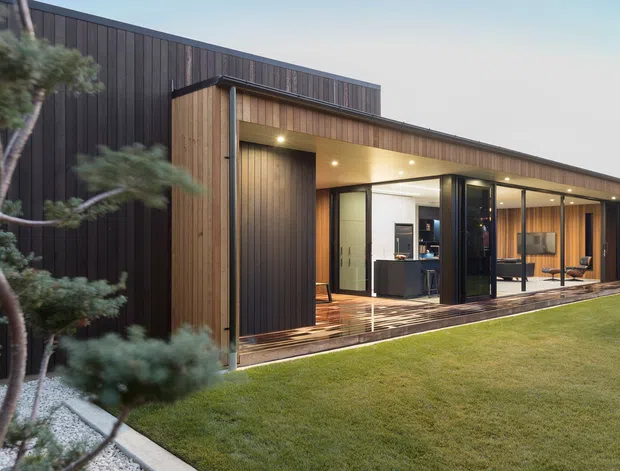 Contemporary single-storey residence viewed from the landscaped garden, with dark vertical cladding, warm timber-lined walls and soffits, extensive floor-to-ceiling glazing, and a covered timber deck opening to an open-plan interior. A compact sculptural tree and gravel bed frame the foreground.