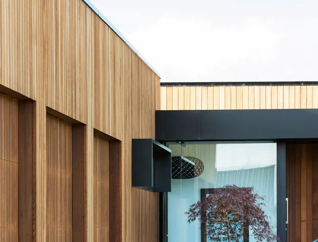 Contemporary building exterior with a strongly geometric composition, featuring warm vertical timber cladding, dark metal trim and soffit elements, large glazed openings, and a pale concrete driveway. A black vehicle is parked in the foreground, partially obscuring the entrance.