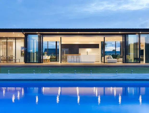 Wide exterior view of a contemporary single-storey residence at dusk, with extensive floor-to-ceiling glazing opening onto a pool. The illuminated interior visibly includes an open-plan kitchen with island and bar seating, a dining area, and a living room. The building has a simple low-pitched roof, dark slender framing, light-toned interior finishes, and a long reflecting swimming pool in the foreground.
