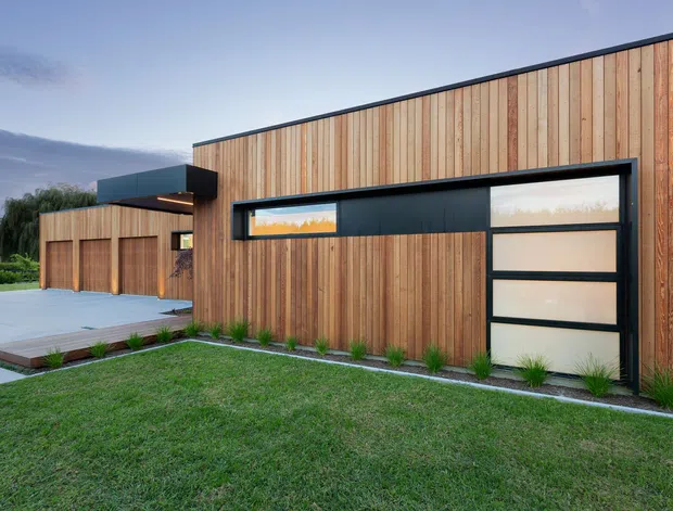 Exterior view of a contemporary single-storey residence or ancillary building clad in vertical timber boards, with a long dark-framed glazed garage door, low flat roofline, timber fencing, concrete paving, and landscaped lawn. The clean rectilinear composition and warm timber facade create a modern, understated appearance.