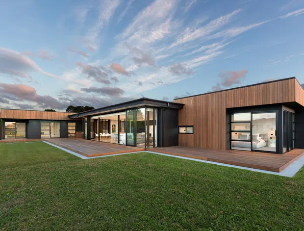 A single-storey contemporary home set in a spacious landscaped lawn, featuring dark metal cladding, warm vertical timber cladding, expansive floor-to-ceiling glazing, and a low-pitched roof. A central glazed living area connects to timber decks, with attached garage or ancillary structures extending to the left. The dusk sky and surrounding greenery create a calm, modern residential setting.