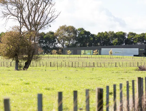 A low, contemporary rural building set within open grassy paddocks. The long dark facade includes large areas of glazing and a lighter grey rectangular volume, with mature trees and simple post-and-wire fencing surrounding the property.