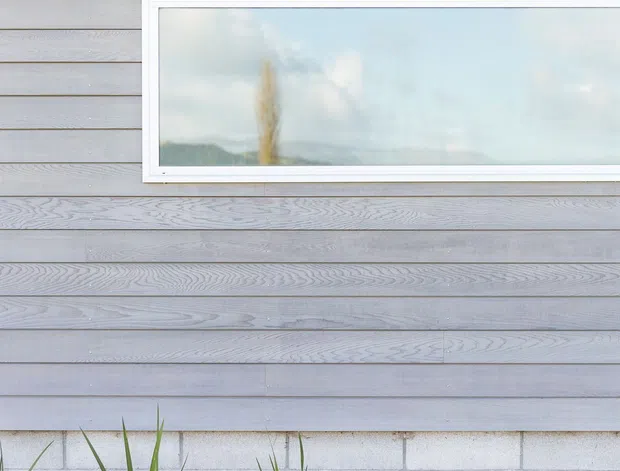 Close-up exterior view of a contemporary building facade with horizontal grey timber weatherboard cladding, a long narrow white-framed window, and a lower base of light concrete masonry blocks. A simple landscaped strip with grasses, mulch, and lawn runs along the wall.