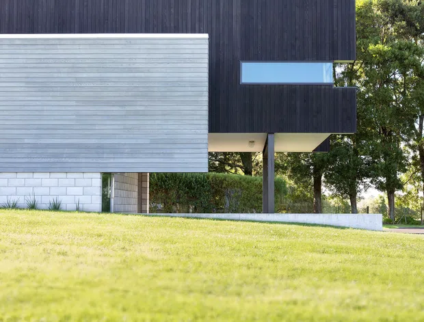 Exterior architectural photograph of a contemporary elevated building set within a landscaped grassy site. The facade combines dark vertical timber cladding with a projecting pale horizontal-clad volume, narrow horizontal glazing, exposed supports, and a low masonry retaining wall.