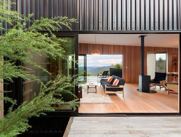 Modern architectural exterior view looking through a large open glazed wall into a warm, timber-lined living room. The building has dark vertical metal cladding, expansive black-framed glazing, a timber deck, and a projecting roof or upper volume. Inside are mid-century-inspired lounge chairs, a central wood-burning stove, timber flooring and walls, and a broad outlook toward a mountainous landscape.
